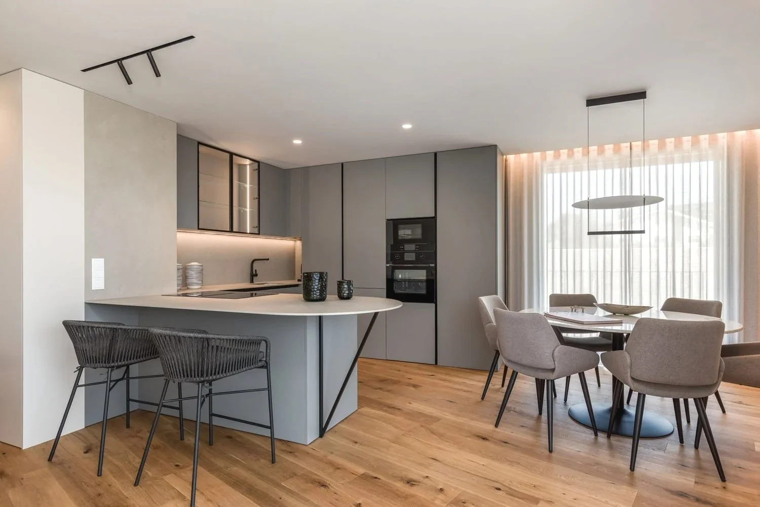 Cozinha moderna integrada à sala de jantar com móveis em tons neutros, piso de madeira clara, iluminação embutida e cortinas de tecido translúcido na janela grande ao fundo.