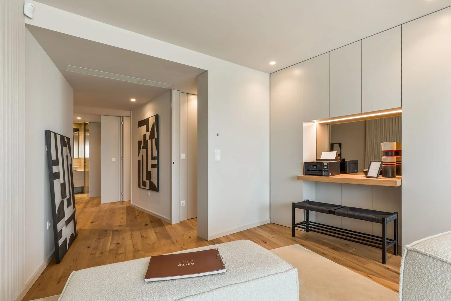 Sala de estar moderna com móveis brancos, piso de madeira, espelho na parede, papel de parede listrado, estante com objetos decorativos, e um espaço de escritório com impressora e prateleiras.