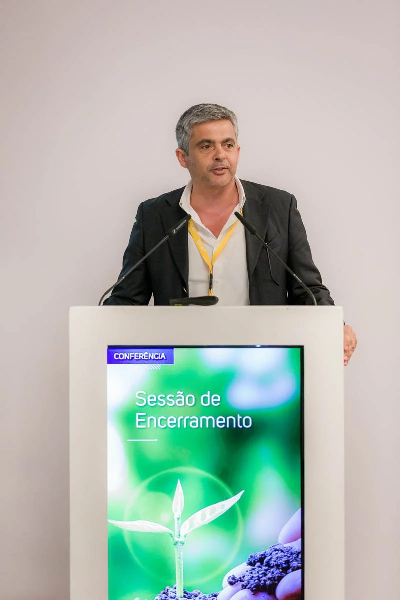 Homem de terno preto e camisa branca falando em um púlpito com tela digital, fundo branco.