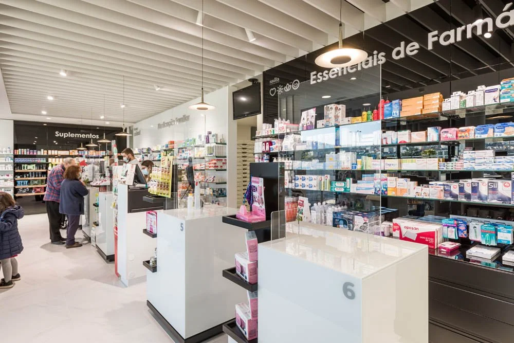 Interior de farmácia com prateleiras cheias de medicamentos e suplementos, atendentes ajudando clientes na caixa, e sinais indicando 'Suplementos' e 'Essenciais de Farmácia'.