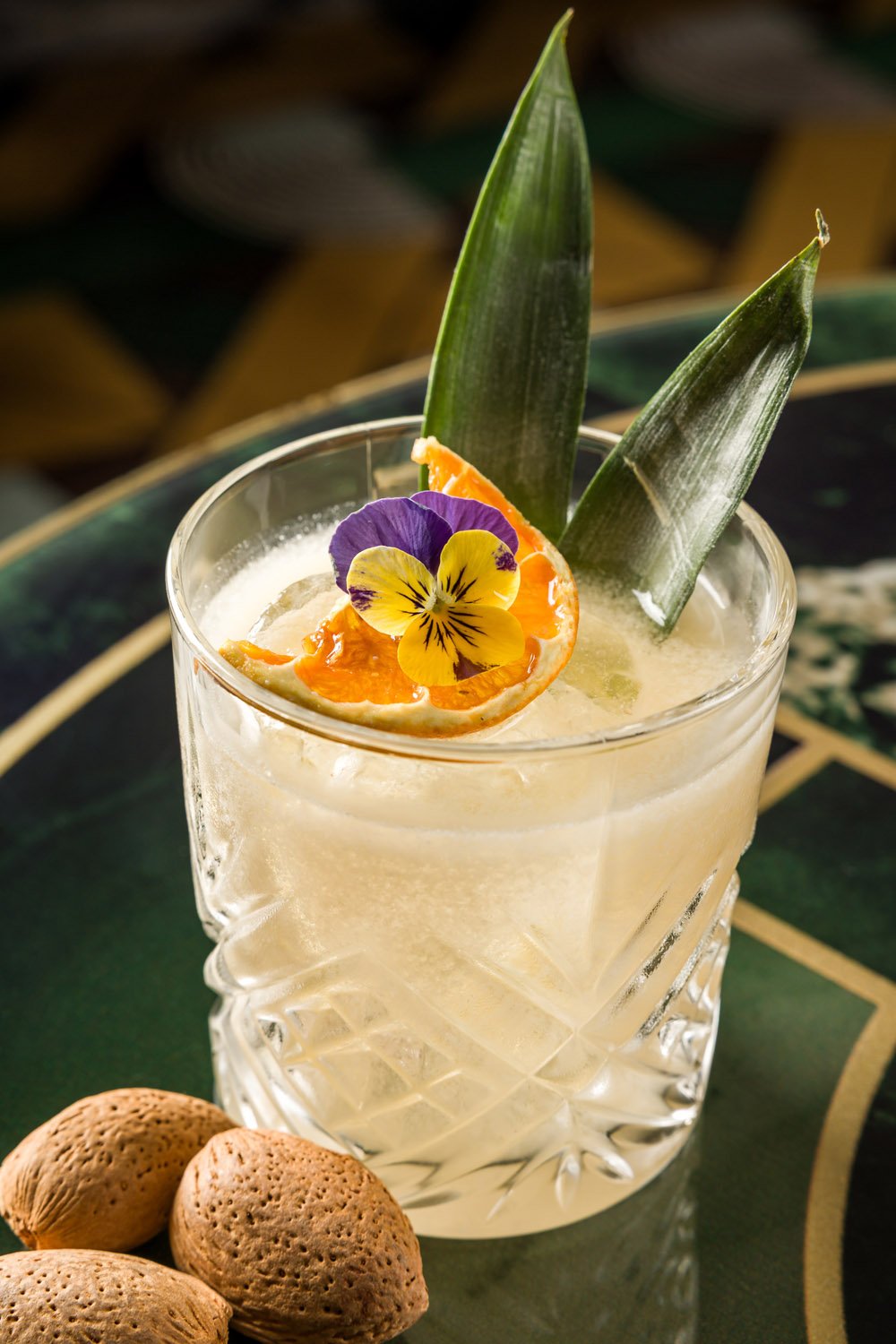 Copo de drink com decoração de abacaxi, flor de viola, fatias de laranja e gelo, sobre mesa de prato verde escura.
