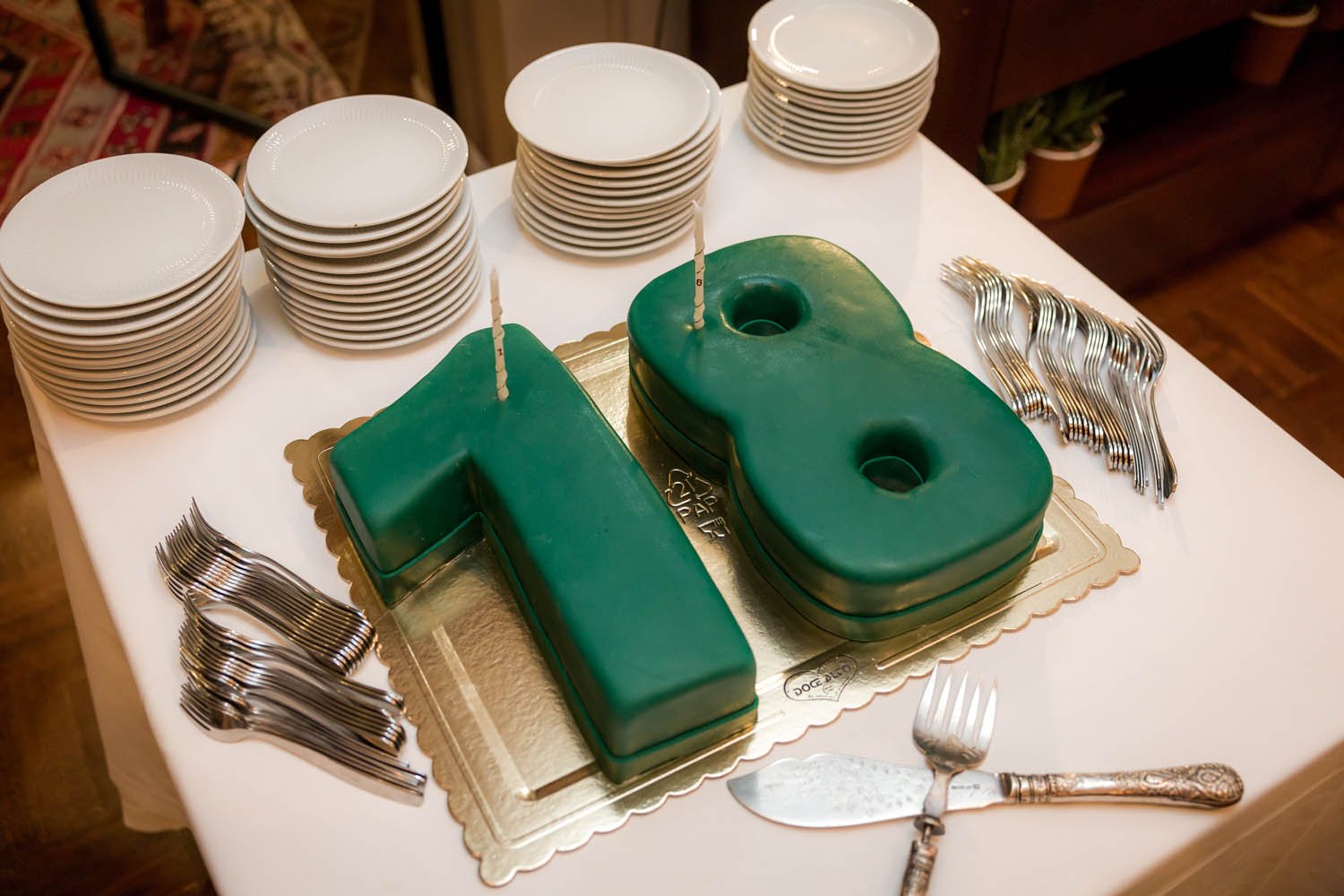 Bolo de aniversário em formato de número 18, decorado com cobertura verde, acompanhado de pratos brancos, garfos e uma faca em uma mesa.