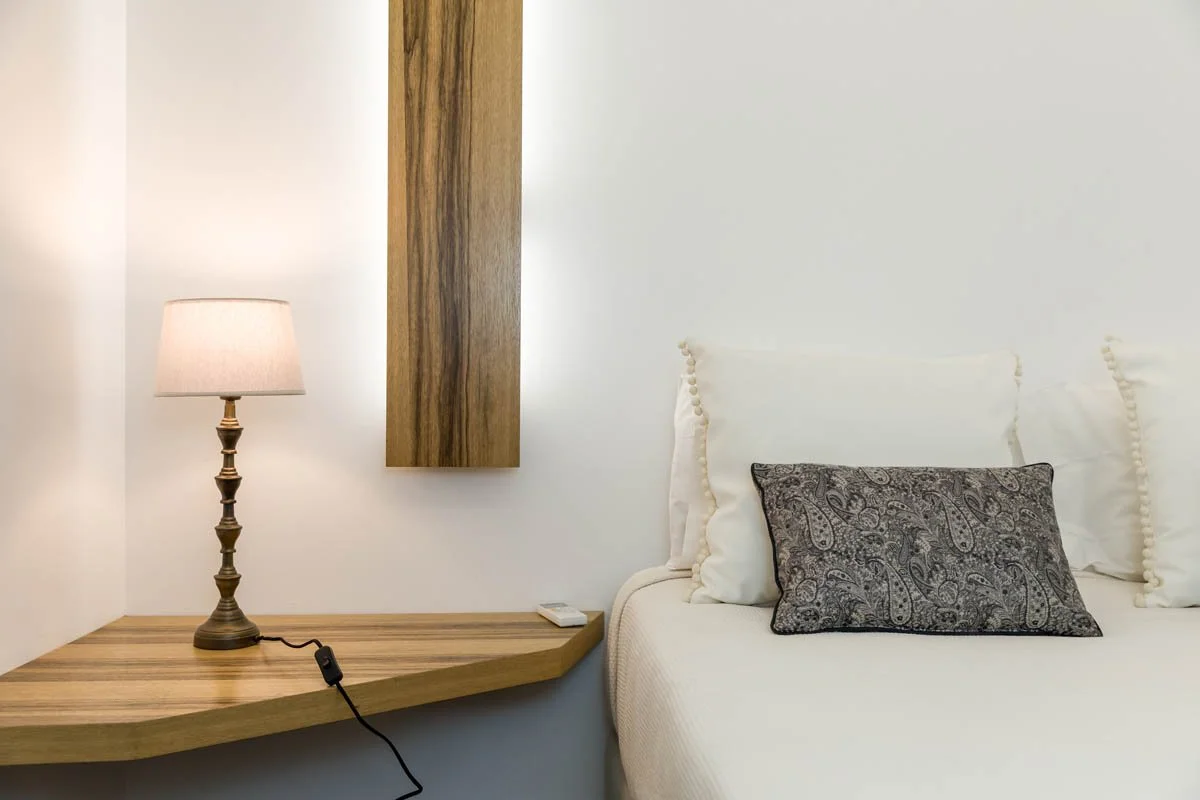 Cama com travesseiro de estampa paisley e cabeceira branca, parede branca decorada com uma moldura de madeira, ao lado uma mesa de cabeceira de madeira com uma luminária de mesa de estilo clássico