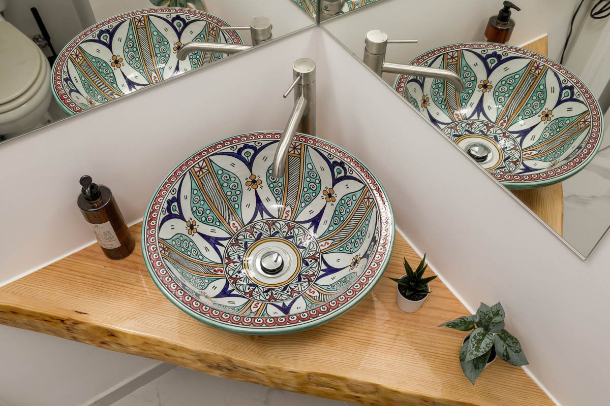 Pia de banheiro com pia de cerâmica decorada com padrão colorido, espelho, sabonete líquido, plantas em vasos e base de madeira.