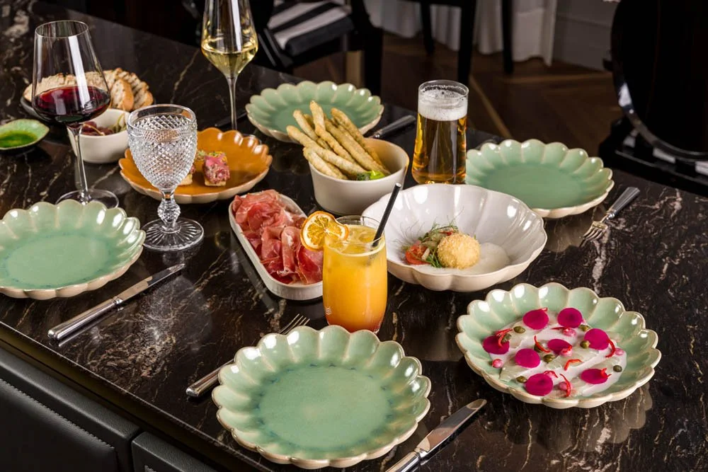 Mesa de jantar com pratos vazios, copos de vinho tinto, água, cerveja, suco de laranja, pão, frias, e petiscos variados, pronta para uma refeição ou celebração.