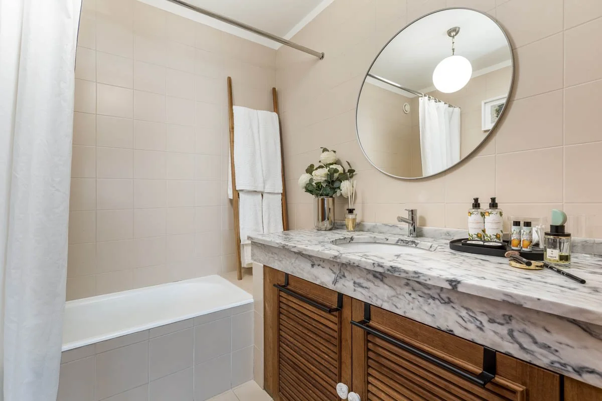 Banheiro com pia de mármore, espelho redondo na parede, prateleira de madeira com toalhas, pia com torneira moderna, decoração com flores, sabonetes e outros itens de higiene, chuveiro com cortina branca, iluminação com luzes de teto.