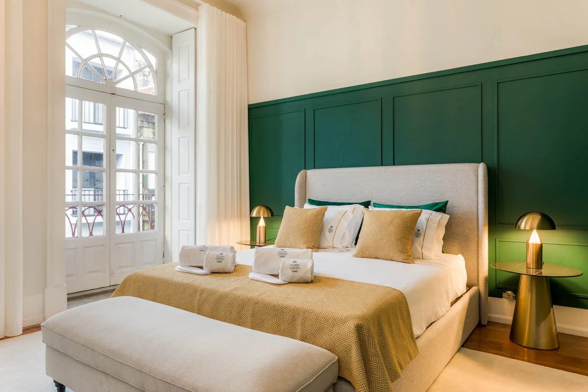 Quarto de hotel com cabeceira de cor cinza claro, paredes verdes e decoração moderna, com roupa de cama branca, almofadas bege e verdes, janelas grandes com cortinas brancas, mesas de cabeceira douradas com luminárias douradas, e uma chaise na frente