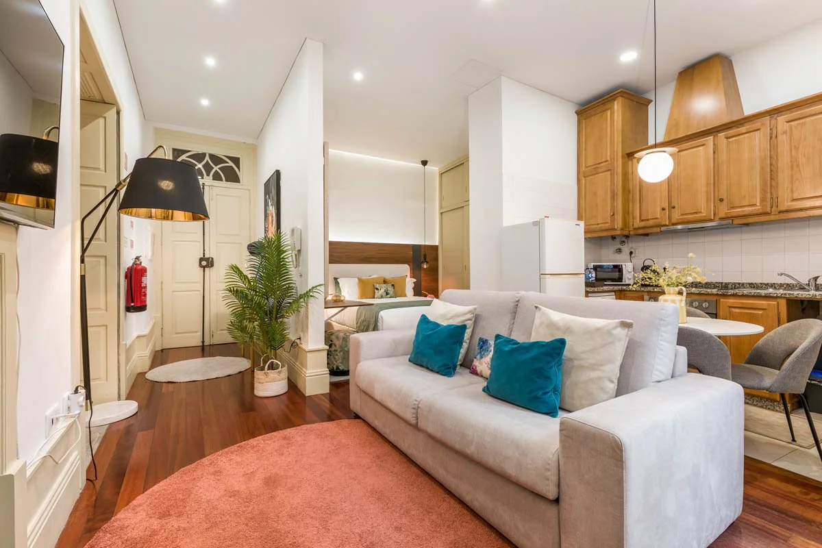 Sala de estar com sofá branco, almofadas azul e bege, tapete coral, cozinha com armários de madeira e geladeira branca, quarto ao fundo com cama e cabeceira de madeira, piso de madeira.