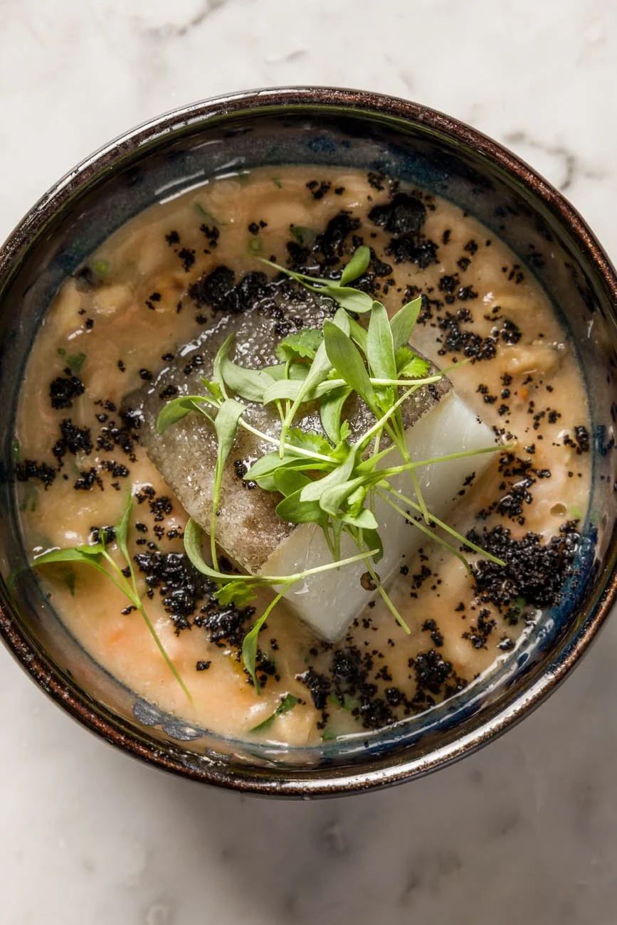 Sopa cremosa com pedaços de peixe, sementes pretas e folhas verdes garnidas por cima.