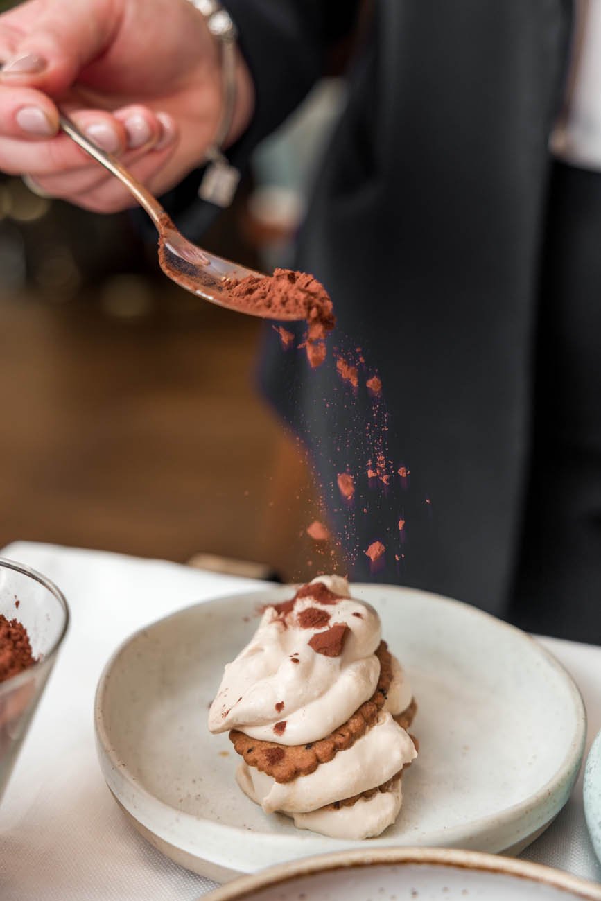 Pessoa polvilhando cacau em pó sobre uma sobremesa com chantilly e biscoitos em um prato