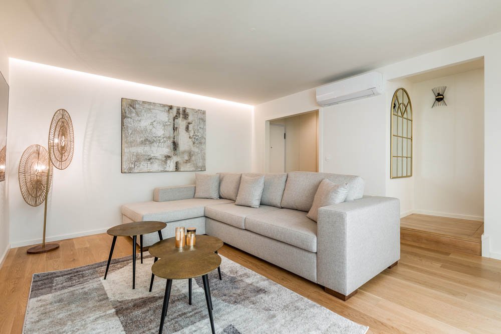 Sala de estar com sofá grande cinza, mesas de centro pequenas, iluminação de piso com três abajures, parede decorada com quadro e espelho, piso de madeira, ar-condicionado split.