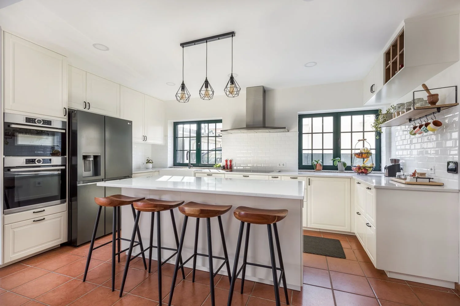 Cozinha moderna com ilha central, iluminação pendente, janelas com moldura verde, armários brancos, piso de cerâmica vermelha, eletrodomésticos de inox, prateleiras decorativas e utensílios de cozinha.