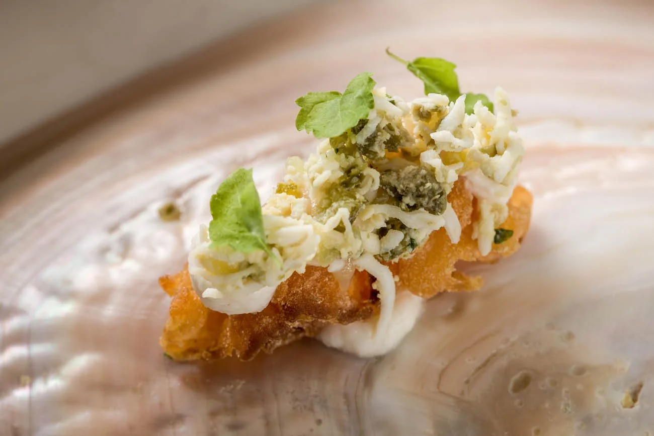 Porção de comida com peixe empanado, coberto com queijo ralado, folhas verdes e molho branco em um prato de pedra