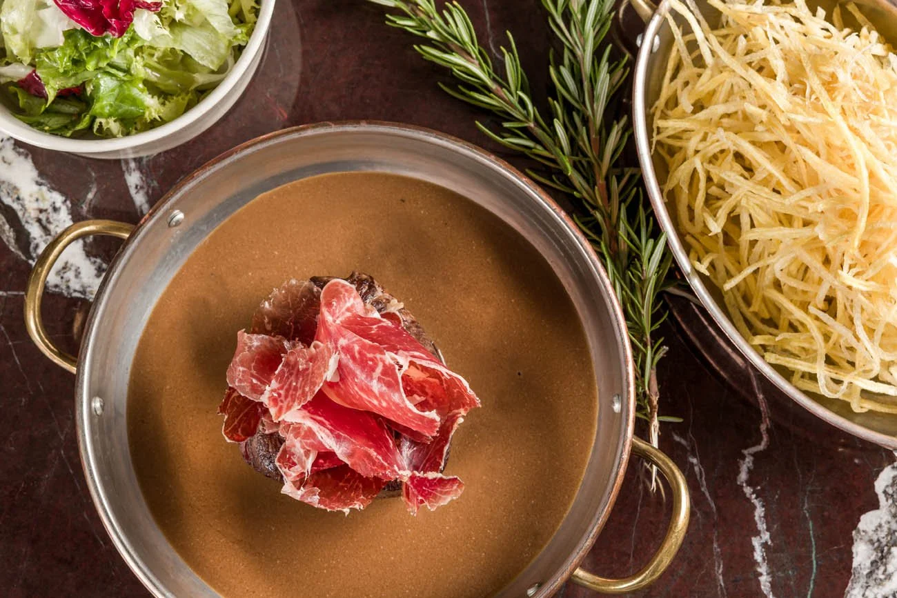 Panela com caldo de carne com pedaços de presunto e carne, ao lado uma tigela com queijo ralado, uma tigela de salada com folhas verdes e um ramo de alecrim.
