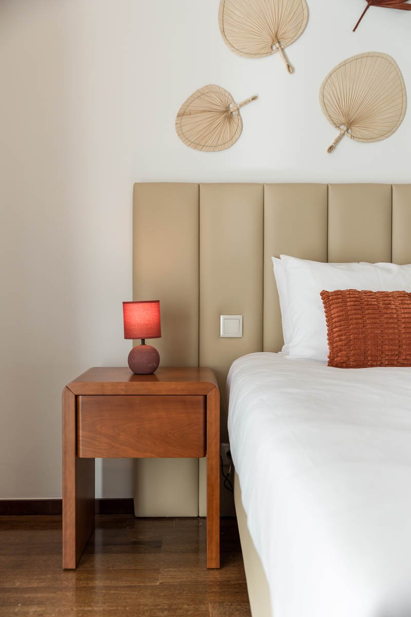 quarto com cabeceira almofadada bege, criado-mudo de madeira com luminária de abajur vermelho, cama com roupa branca e travesseiro laranja, decoração de parede com cinco leques de palha.