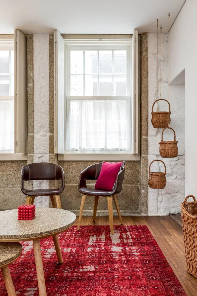 Sala de estar com duas poltronas de couro marrom, uma com almofada rosa, mesa de centro de madeira e duas janelas grandes com cortinas brancas. Há uma parede de pedra e um tapete vermelho, além de prateleiras com cestas de vime penduradas na parede.