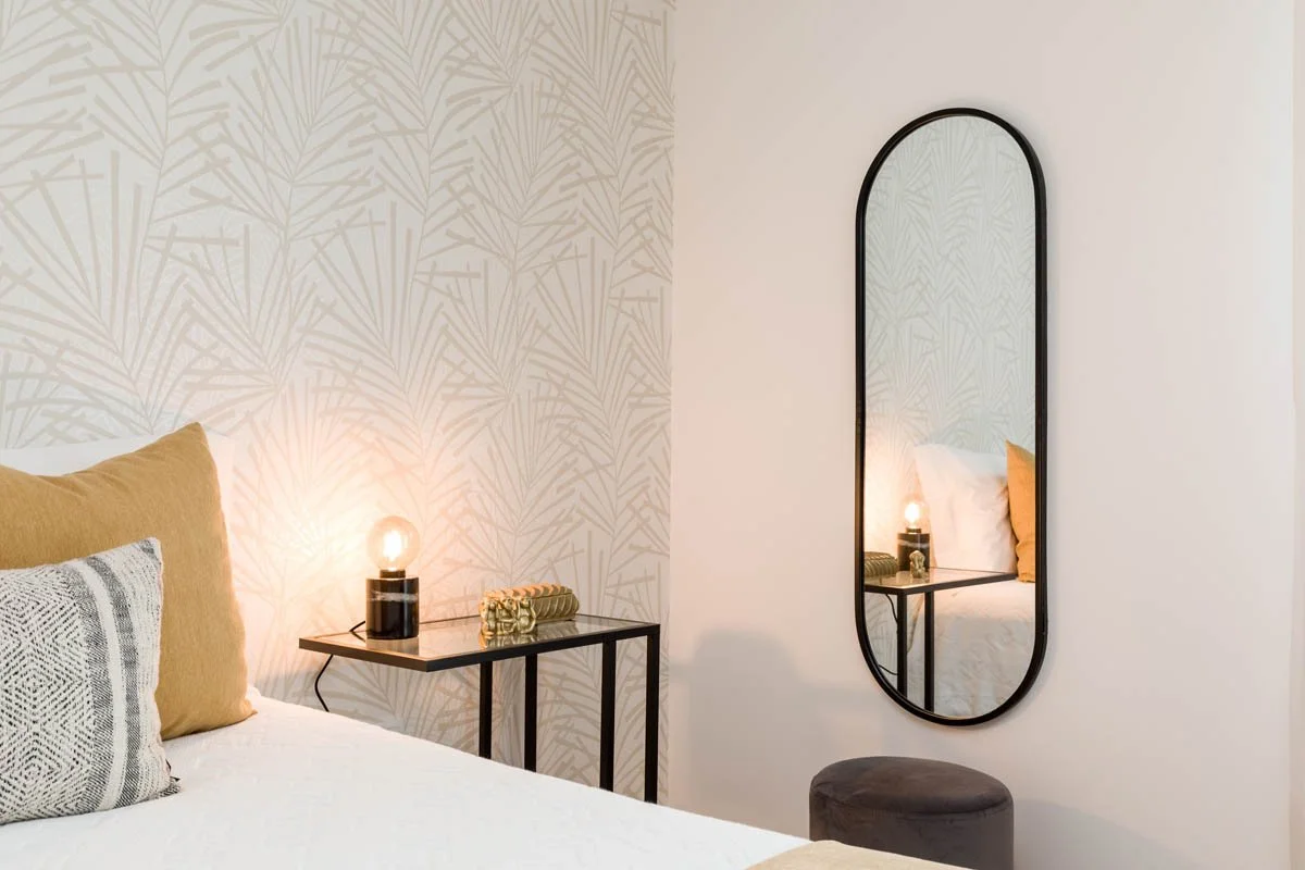 Quarto com parede de papel de parede com padrão de folhas, cama com travesseiro amarelo e outro de padrões gráficos, luminária ao lado da cama, espelho de corpo inteiro com moldura preta e um pufe cinza escuro.