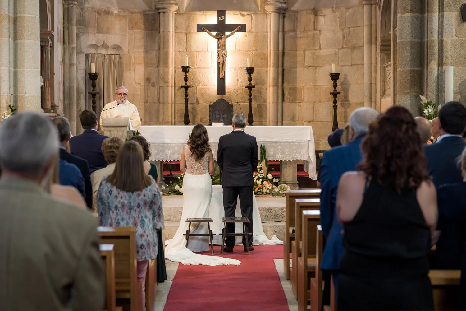 Casamento religioso dentro de uma igreja com o padre, no altar, e um casal se despedindo na frente, com várias pessoas assistindo e uma cruz com Jesus ao fundo.