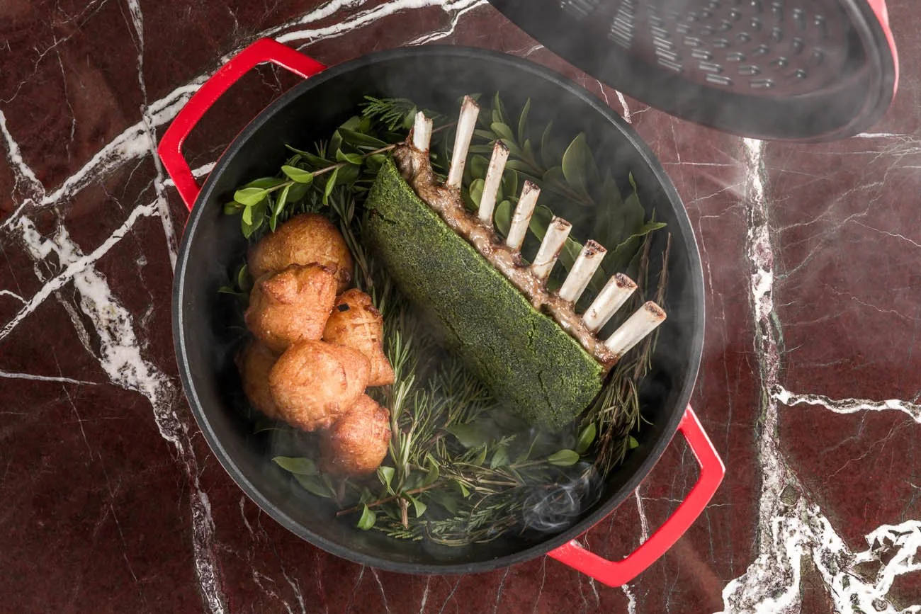 Panela com água fervente, folhas verdes, uma peça de osso de cordeiro, e bolinhos fritos, tudo em um fogão com tampa aberta.