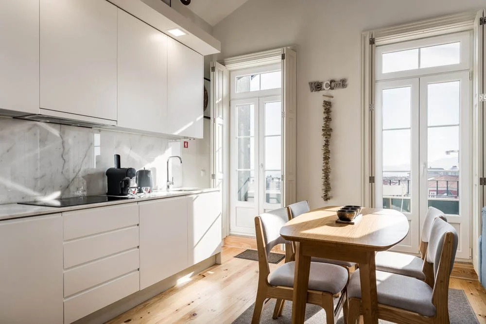 Cozinha moderna com armários brancos, bancada de mármore, mesa de jantar de madeira com quatro cadeiras estofadas, janelas grandes com portas de vidro, decorativa na parede.