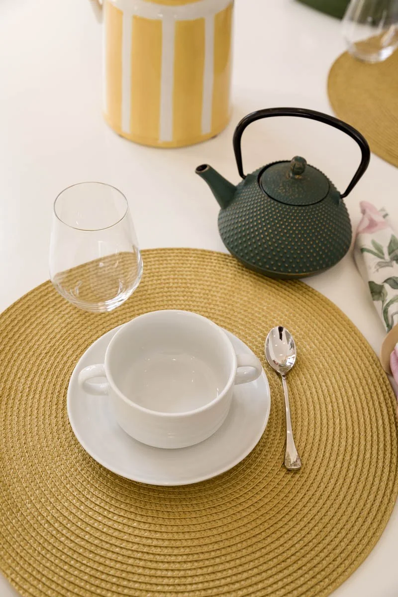 Mesa posta com um prato de fibra natural, uma xícara branca, uma colher de sobremesa, um copo de vidro, uma chaleira verde, um recipiente com latas amarelas e brancas, e uma toalha de pano com estampa floral parcialmente visível.