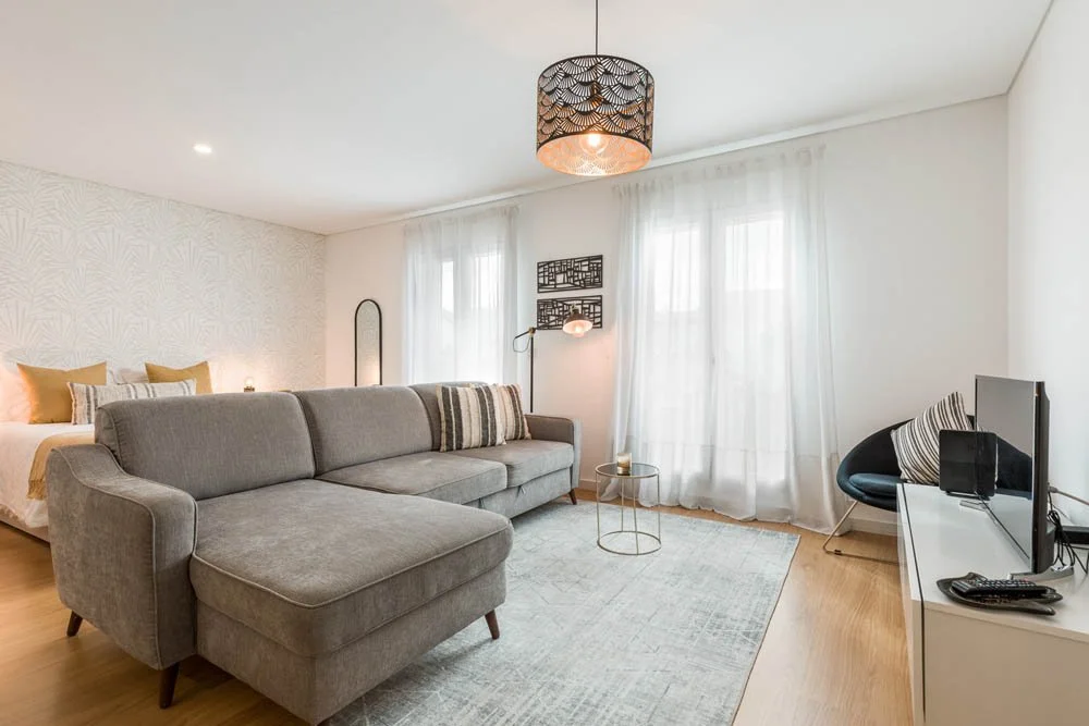 Sala de estar com sofá cinza, TV em um suporte branco, cadeira preta com almofada listrada, mesa de centro pequena com luminária, cortinas leves brancas, parede com decoração moderna, piso de madeira claro e iluminação suave.