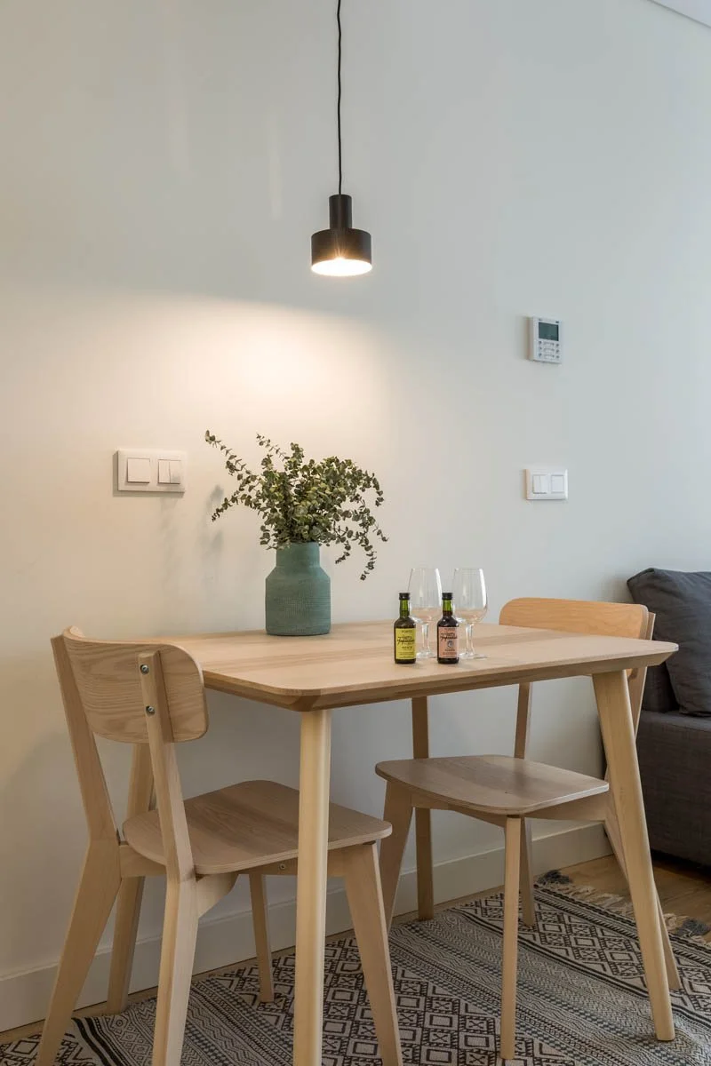 Mesa de jantar de madeira com duas cadeiras de madeira, um vaso com planta e duas taças de vinho, com uma luminária suspensa acima e parede clara ao fundo.