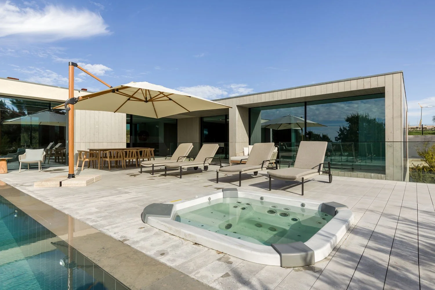 Varanda de casa moderna com espreguiçadeiras, mesa de jantar, guarda-sol, jacuzzi e vista para o céu claro.
