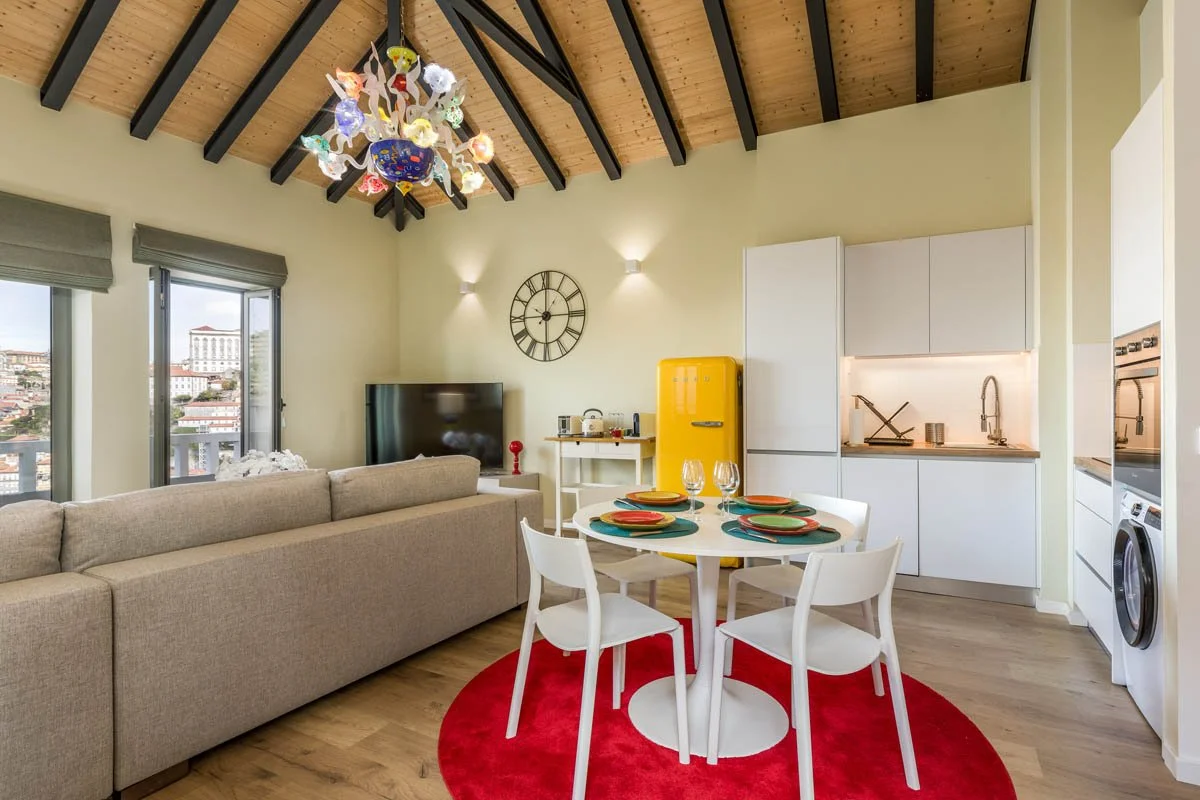 Sala de estar com teto de madeira, chandelier colorido, sofá bege, mesa de jantar com quatro cadeiras brancas, pratos coloridos, taças, parede com relógio grande, televisão, geladeira amarela, cozinha moderna com armários brancos, bancada com pia, el