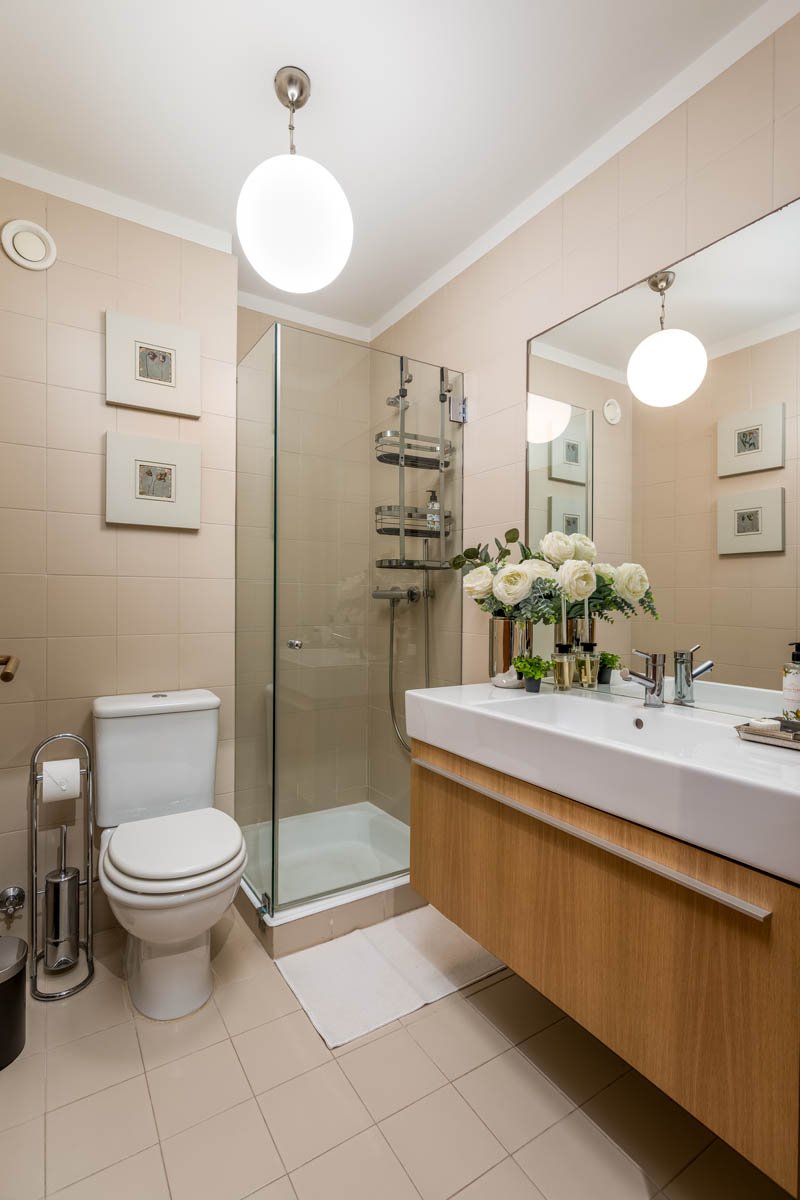 Banheiro moderno com vaso sanitário, box de vidro com chuveiro, pia com espelho grande e decoração com flores brancas