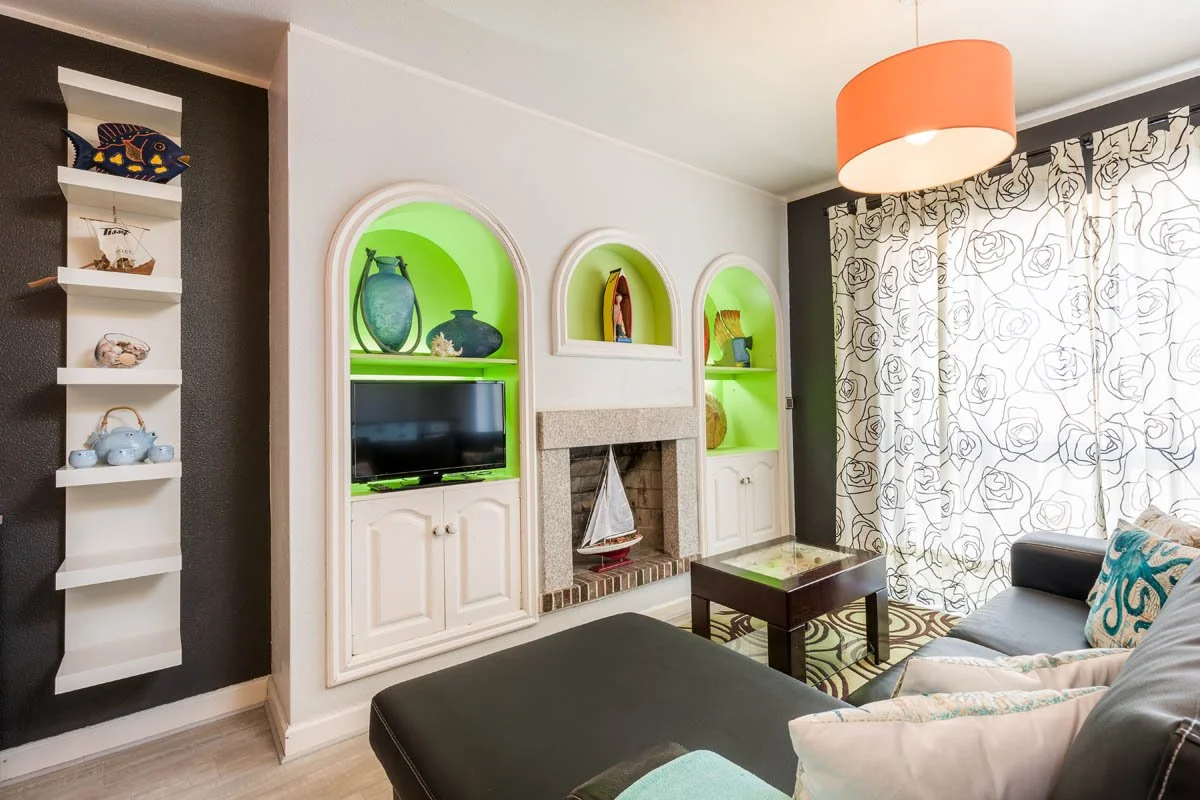 Sala de estar com parede branca, detalhes em preto, estantes brancas com objetos decorativos, lareira decorativa, cortinas com estampa de rosas, sofá cinza com almofadas coloridas, mesa de centro escura, iluminação de teto com luminária cor de coral.