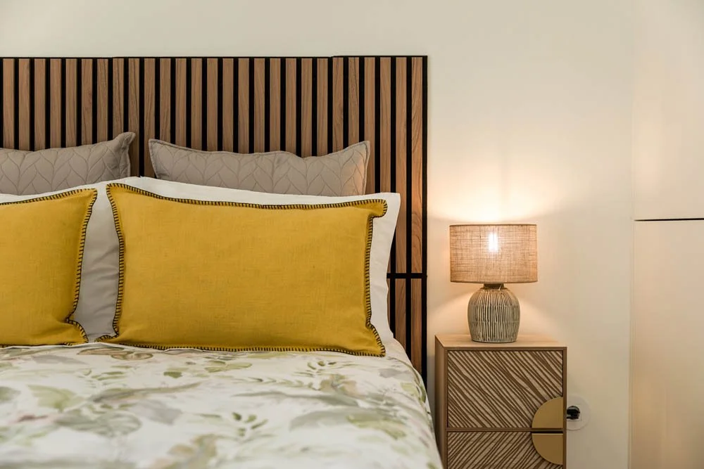Cama com cabeceira de madeira, travesseiros cinza e almofadas amarelas, abajur de mesa ao lado, parede branca ao fundo.