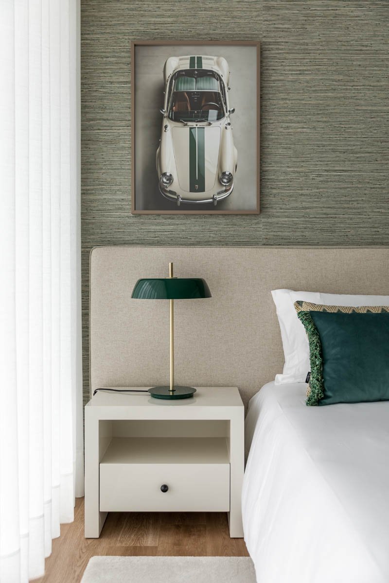 Quarto com parede texturizada, cabeceira bege, mesa de cabeceira branca com gaveta, luminária verde, quadro com imagem de carro esportivo clássico, travesseiro verde e roupa de cama branca.
