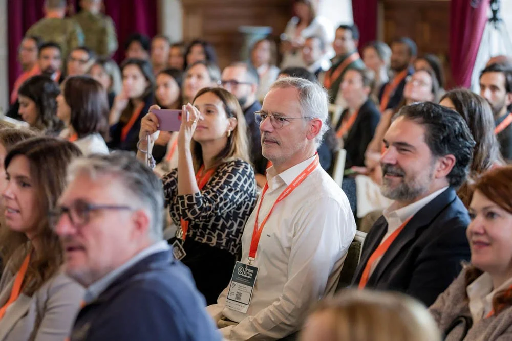 Pessoas assistindo a uma palestra ou conferência em uma sala com esteira de cadeiras, com alguns usando crachás laranjas.