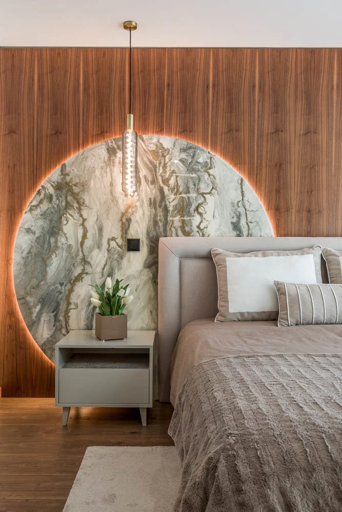 Quarto com parede de madeira e uma parede de mármore com iluminação de LED ao redor de um círculo. Ao lado da cama há uma mesa de cabeceira branca com uma planta de tulipas brancas em vaso rectangular. A cama tem cabeceira acolchoada e está coberta com roupa de cama bege e travesseiros brancos com detalhes suaves.
