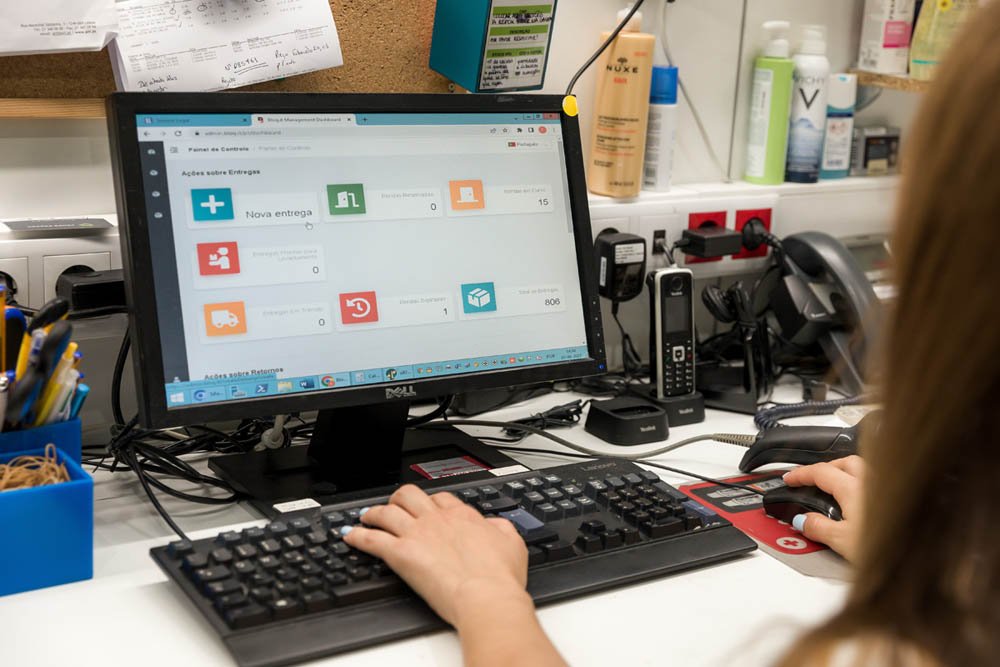 Cena de uma pessoa usando um computador de mesa em um escritório, com a tela exibindo uma interface de sistema de gerenciamento de entregas. Ao redor, estão dispositivos de telefone, uma caneta e materiais de escritório.