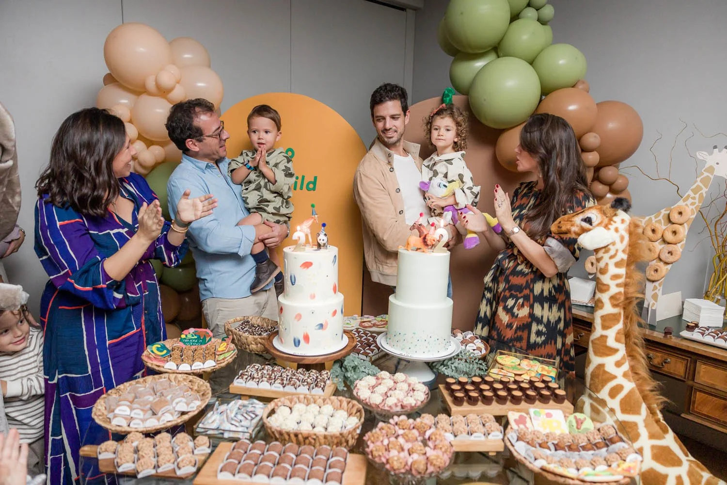 Celebração de aniversário com bolo, decoração de balões e pessoas comemorando com crianças, incluindo um leão de pelúcia ao lado de uma girafa de balão.