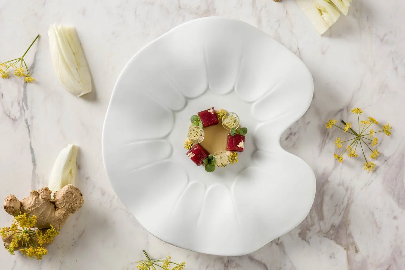 Prato de gastronomia apresentado em um prato branco com formas decorativas, com pequenos pedaços de carne vermelha, fatias de queijo, molho ou caldo e decoração com ervas e flores pequenas ao redor, sobre uma superfície de mármore.