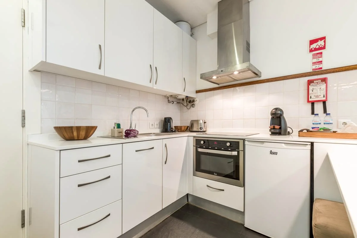 Cozinha compacta com armários brancos, torneira, pia, forno, geladeira pequena, chaleira, torradeira, máquina de café, exaustor e alguns utensílios sobre a bancada.