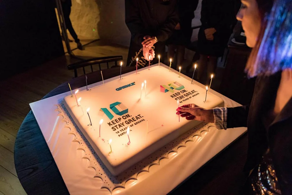 Pessoas ao redor de um bolo de aniversário com velas acesas, uma pessoa está acendendo uma vela, no bolo há texto em inglês que diz 'Keep on, stay great' e números comemorativos 10, indicando uma celebração de aniversário de 10 anos.