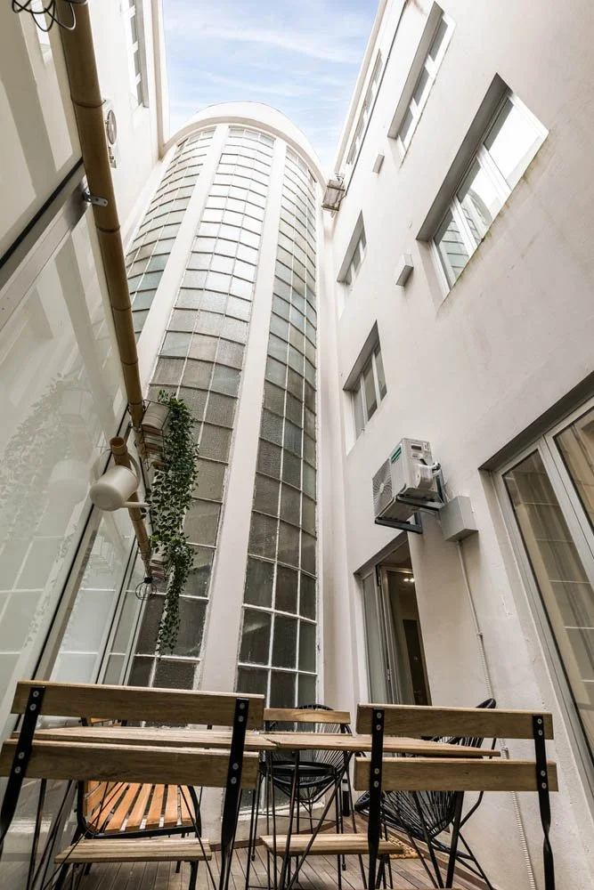 Pátio com mesa de madeira e cadeiras, cercado por paredes brancas de edifícios com janelas e uma parede de vidro em forma de tubo que sobe até o teto, céu visível ao fundo.