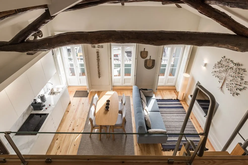 Visão aérea de uma sala de estar moderna com espaço de cozinha integrado, mesa de jantar, sofá, televisão, tapete e decorções em parede, com janelas grandes deixando entrar muita luz natural.