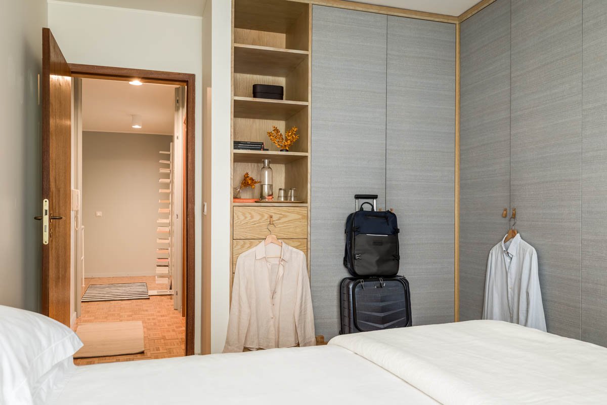 Quarto com armário, roupa pendurada, malas e mochila, vista de uma cama de casal e entrada para o corredor.