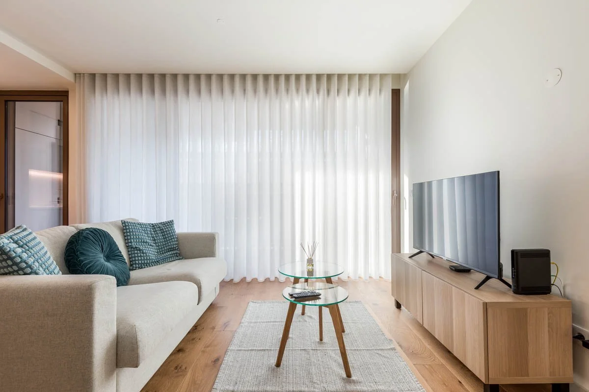 Sala de estar moderna com sofá bege, almofadas azul, mesa de vidro, televisão grande em móvel de madeira e cortinas brancas.