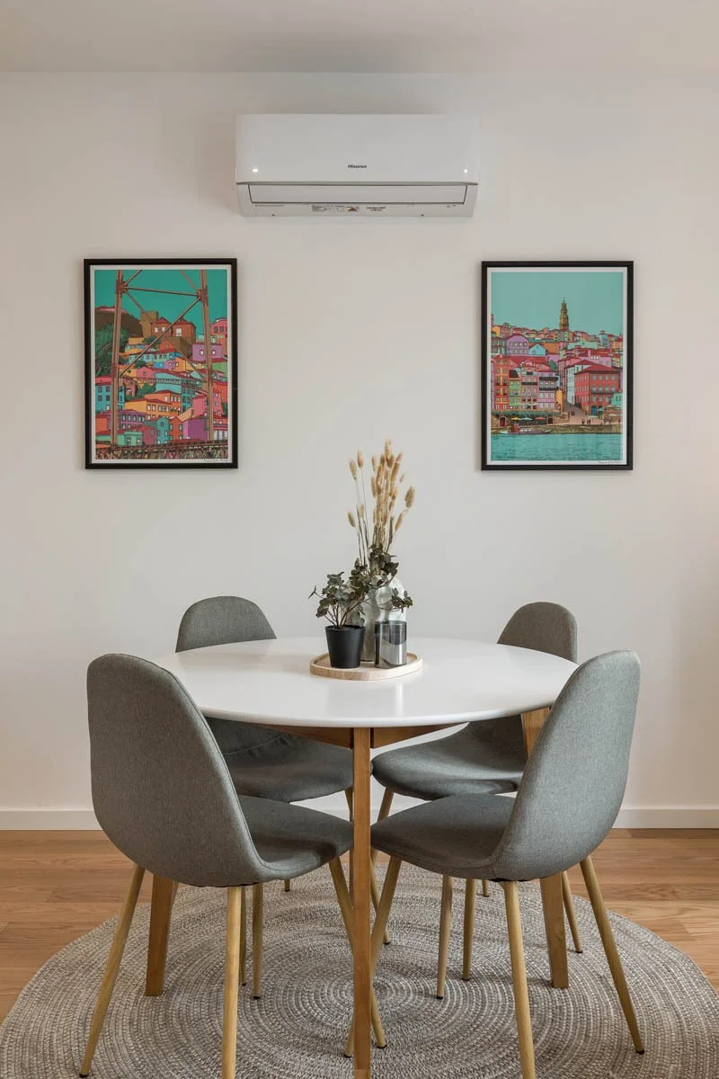 Sala de jantar com mesa redonda branca, cinco cadeiras cinzas, vaso com plantas secas no centro, duas gravuras de paisagens urbanas na parede e ar condicionado acima.