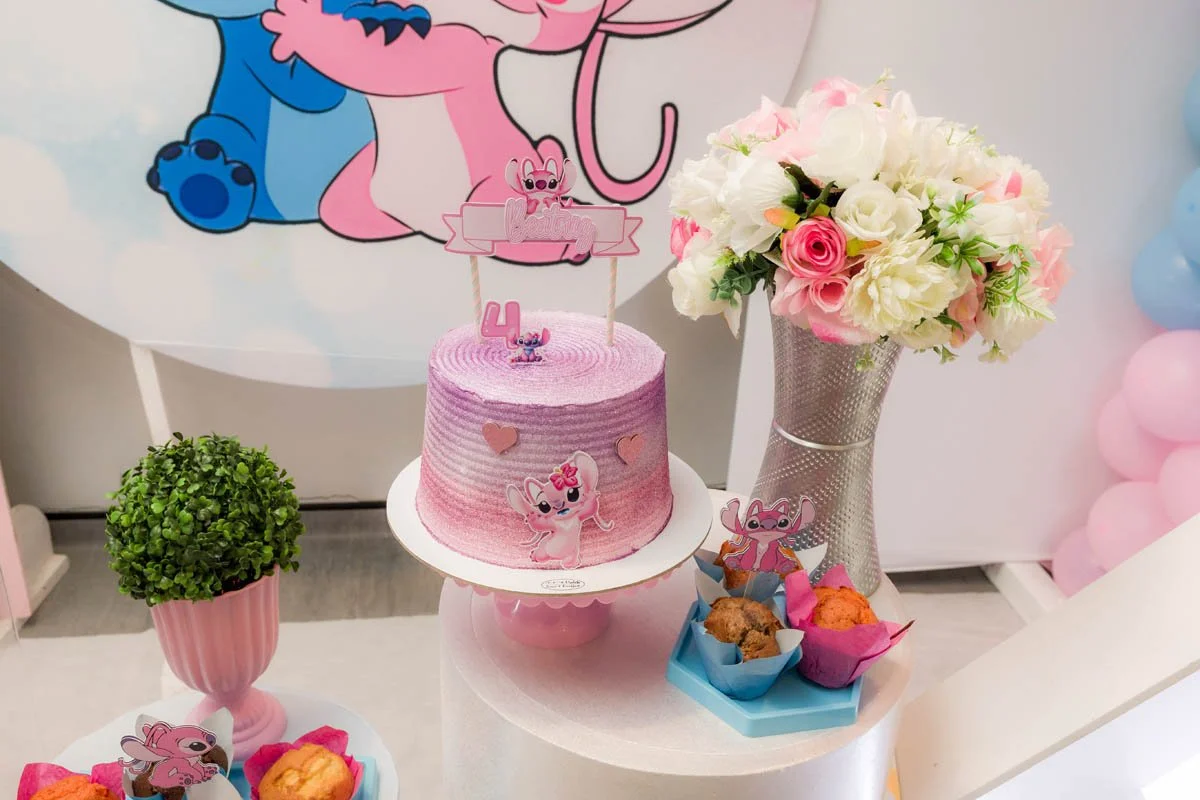 Mesa decorada com tema de aniversário da personagem Biruta, com bolo rosa decorado com personagens, flores em vaso, cupcakes e uma parede com balões coloridos ao fundo.