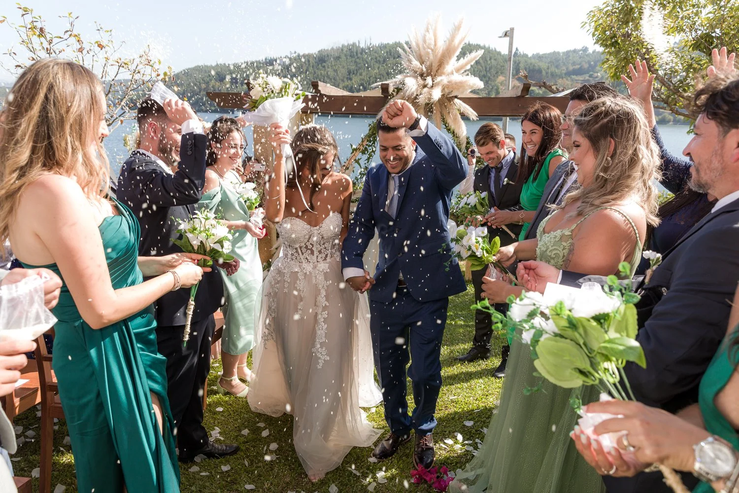 Casamento ao ar livre com o casal de noivos sorridentes e convidados jogando arroz ou pétalas de flores, com cenário de natureza, árvores e água ao fundo.