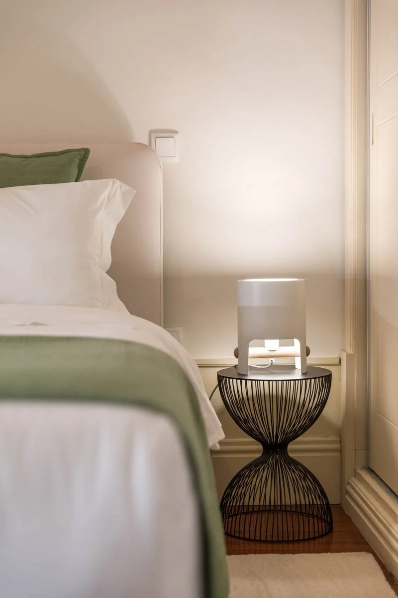 Quarto com cama de cabeceira branca, almofada verde e roupa de cama branca. Ao lado, uma mesa de cabeceira de metal preta com um abajur branco moderno.