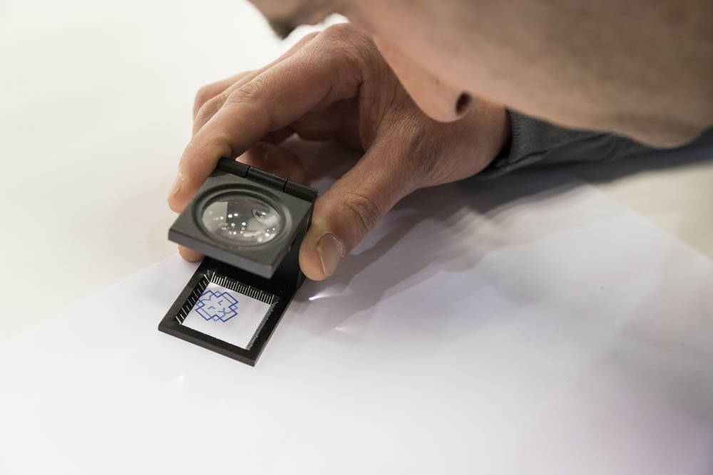 Pessoa usando uma lente de aumento para examinar uma assinatura ou desenho em uma folha de papel.