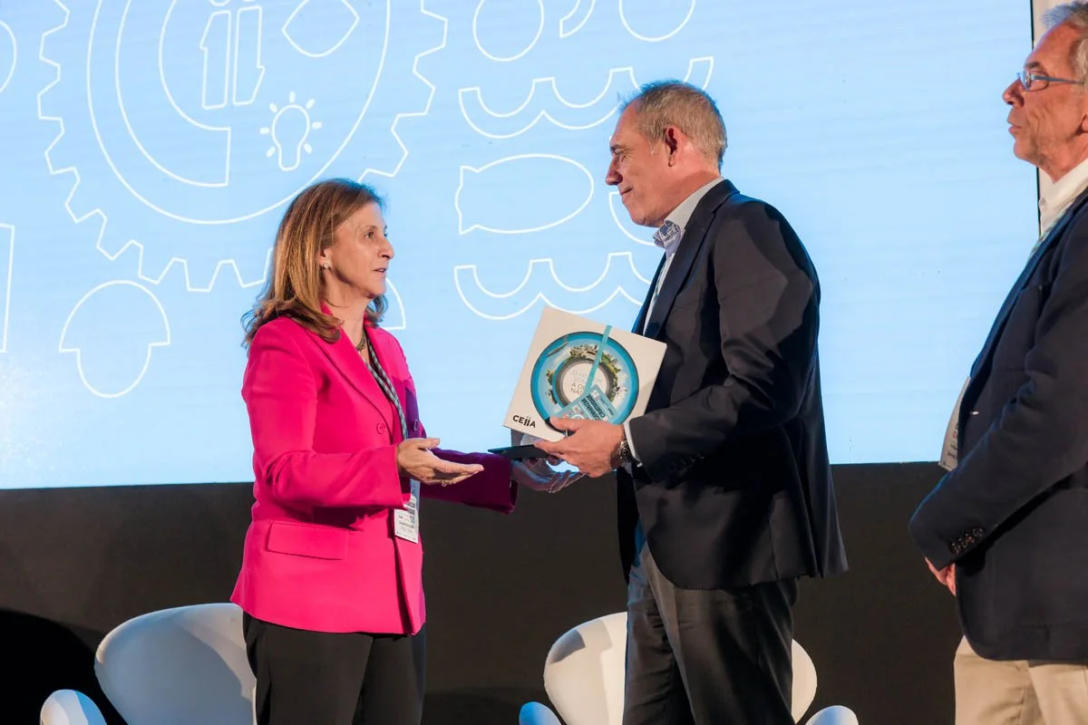 Mulher em blazer rosa recebendo um prêmio de homem com terno escuro em palco durante conferência.